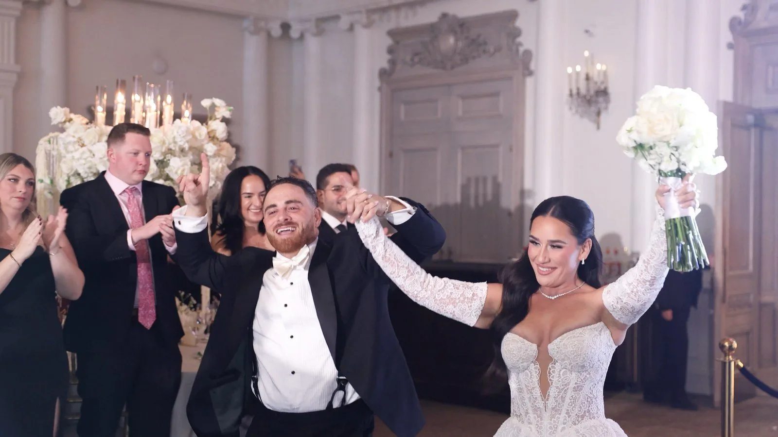 Newlyweds entering their reception at Park Chateau Estate and Gardens, hands raised and bouquet high, as guests applaud in the chandelier-lit ballroom